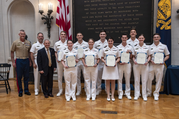 Gruppe uniformierter Personen, die strahlen und Zertifikate halten, mit einem Tisch, der mit blauer Stoffdecke, Stühlen, Fahnen, Lichtern und einer Tafel mit der Aufschrift "U.S. Naval Academy Class of 2018" im Hintergrund geschmückt ist.
