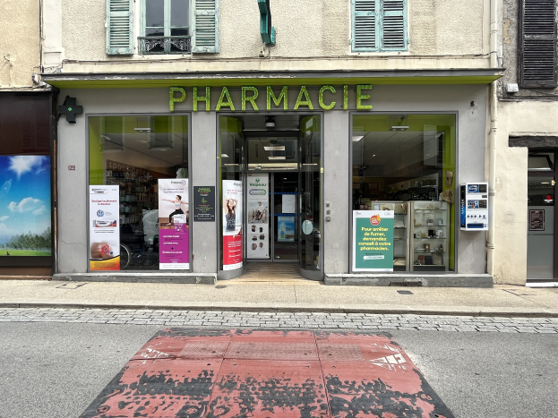 Apotheke mit Glaswänden und -türen, Plakate an den Wänden, an der Ecke einer Straße mit einer Straße im Vordergrund.