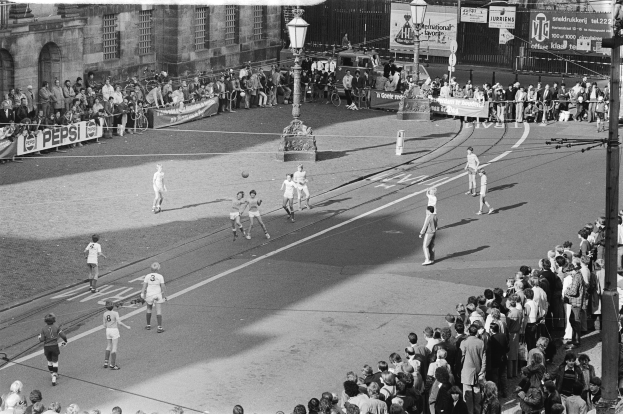 Schwarzes Bild eines Street-Soccer-Spiels mit Spielern und Zuschauern, umgeben von Laternenmasten, Gebäuden und Bannern.