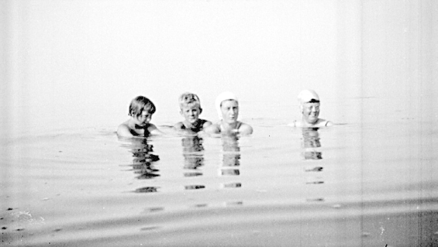 Schwarzes und weißes Foto von vier Kindern, die in einem Pool schwimmen, jedes trägt eine Schwimmkappe.