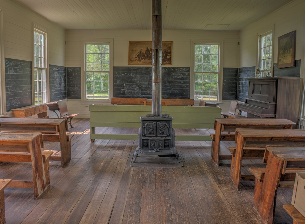 Leerer Klassenraum mit hölzernen Tischen und Stühlen in Reihen, eine mittlere Säule, ein Piano auf der rechten Seite und Fenster mit Bäumen im Freien.