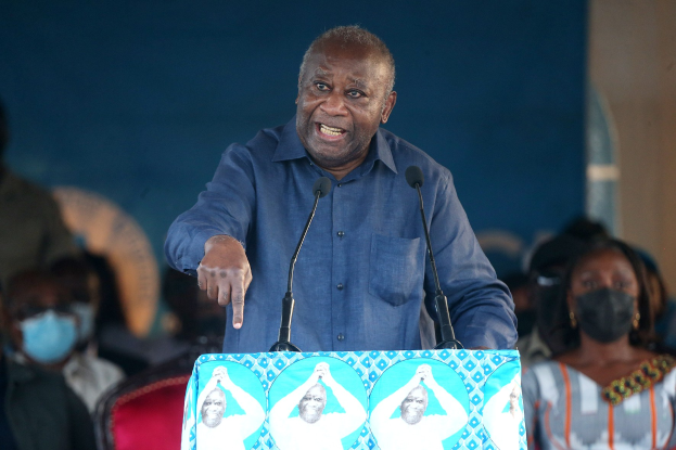 Ein Mann in einem blauen Hemd spricht in zwei Mikrofone an einem Rednerpult, hinter ihm sitzt eine Menge, einige tragen Masken, und es ist eine Wand im Hintergrund zu sehen.