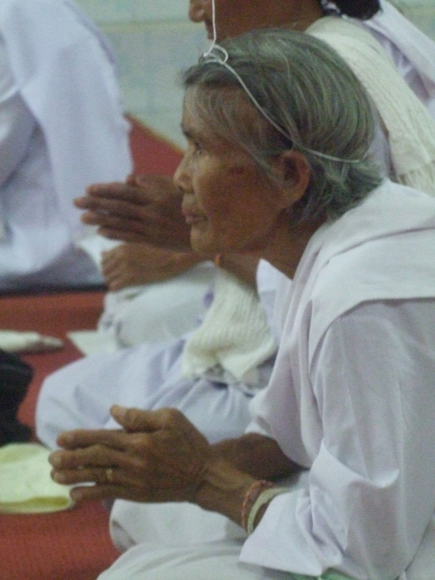 Eine ältere Frau liegt in einem Krankenhausbett und hat ihre Hände gefaltet, sieht besorgt aus mit geschlossenen Augen und Kopf auf den Händen ruhend, umgeben von ein paar Menschen im Hintergrund.