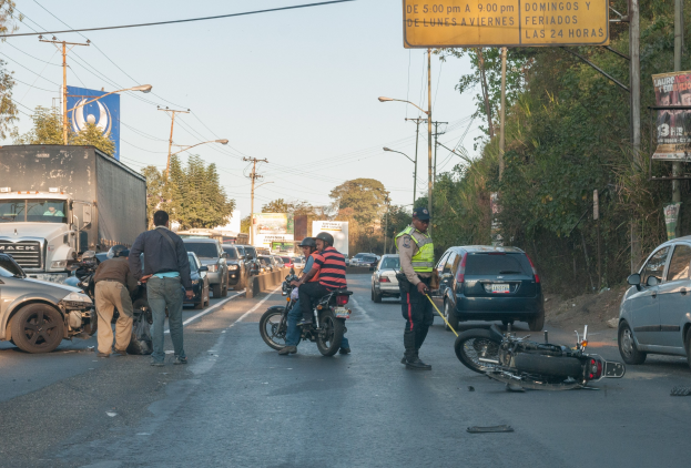 Gruppe von Menschen um ein verunglücktes Motorrad auf dem Seitenstreifen mit mehreren Fahrzeugen, Bäumen und Strommasten im Hintergrund.