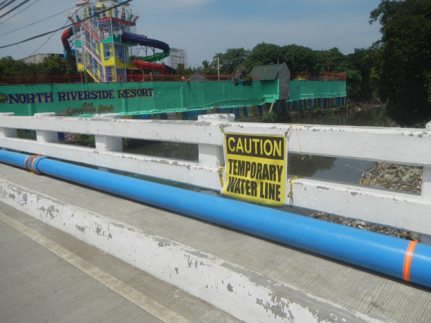 Vorsichtsschild mit der Aufschrift "Vorsicht temporäre Wasserleitung" auf einer Brücke mit Geländer, Bäumen, Gebäuden, einer Wasserrutsche und einem bewölktem Himmel im Hintergrund.