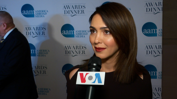 Emmy Rossum in einem schwarzen Kleid hält ein Mikrofon und steht neben einem Mann mit einer Tafel, auf der "Aam Awards Dinner" steht.