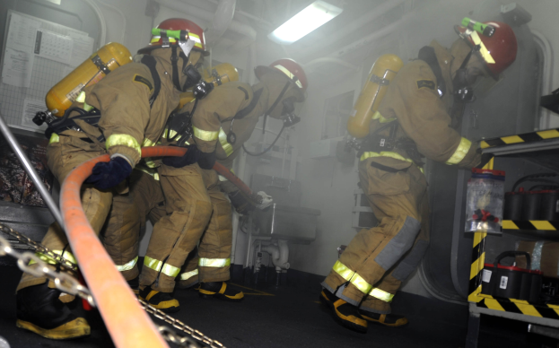 Feuerwehrleute in Schutzausrüstung mit Helmen und Sauerstoffflaschen arbeiten an einem Feuerwehrauto, mit einem Gestell voller Gegenstände und im Hintergrund sichtbare Rohre und Lichter.