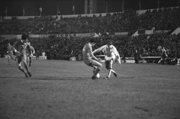 Schwarzes und weißes Bild von Männern, die Fußball auf einem Feld in einem Stadion mit Zuschauern in den Rängen und Fahnen im Hintergrund spielen.