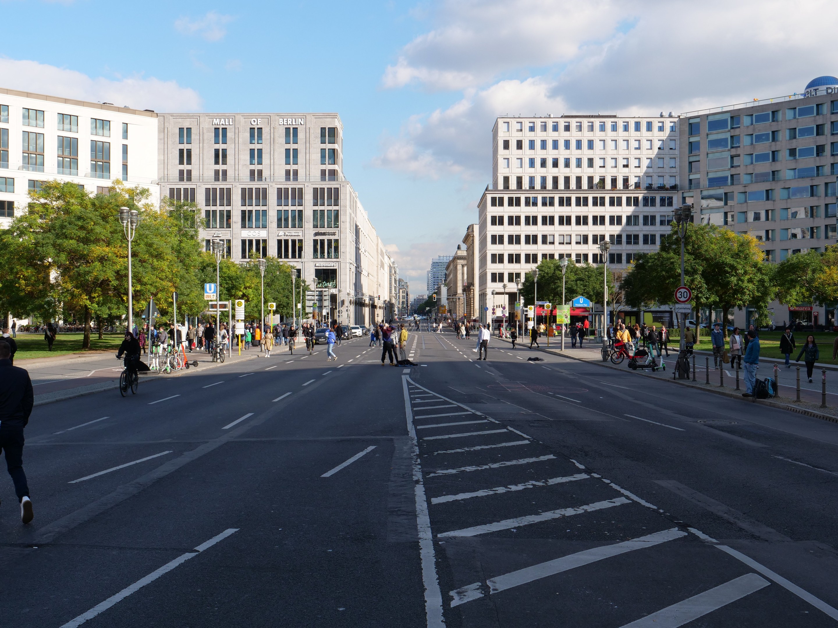 Eine belebte Stadtstraße in Berlin, Deutschland, mit Fußgängern und Radfahrern auf der Straße, hohen Gebäuden, Bäumen, Laternen und Schildern unter einem bewölkten Himmel.