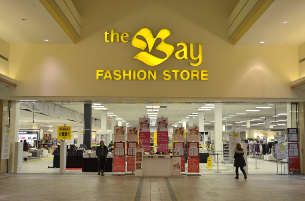 Außenansicht eines Bay Fashion Stores in einem Einkaufszentrum mit Einkäufern, Wandtexttafeln, Säulen, Deckenlampen und Hintergrundauslagen.