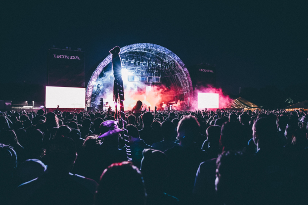 Menschenmenge auf einem Musikfestival mit einem Künstler auf der Bühne umgeben von Musikern, beleuchtet von Scheinwerfern und Bildschirmen.