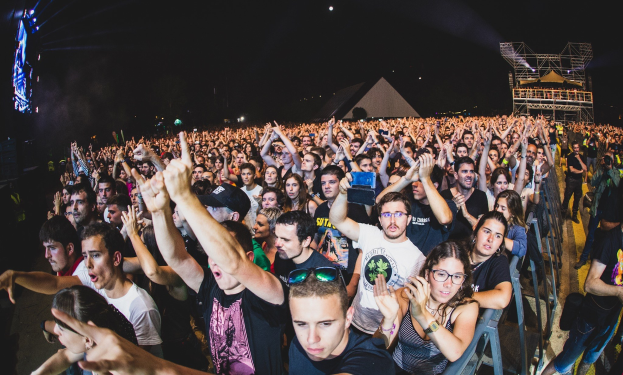 Eine große Menschenmenge auf einem Musikfestival mit Menschen, die ihre Hände heben, einige sitzen und andere stehen, mit einem Metallzaun auf der rechten Seite, Gebäuden und Lichtern im Hintergrund und einem dunklen Himmel.