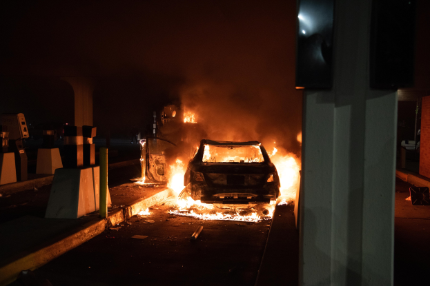 Ein Auto in Flammen an der Straße bei Nacht, mit einer Säule auf der rechten Seite und verstreuten Gegenständen auf der linken Seite, in der Nähe einer Tankstelle.