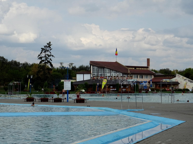 Ein großes Schwimmbad mit Menschen darin, umgeben von Pfählen, Bänken, Topfpflanzen, einem Schild, einem Fahnenmast mit Flagge, einem Gebäude mit Fenstern, Straßenlaternen, einer Baumgruppe und einem bewölkten Himmel.