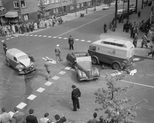 Schwarzes und weißes Bild eines Autounfalls an einer Straßenkreuzung mit mehreren Fahrzeugen, darunter ein Van, und einer Gruppe von Schaulustigen; Gebäude, Bäume, Pfosten und Schilder sind im Hintergrund zu sehen.