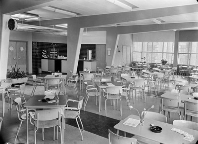 Schwarzes und weißes Foto einer Cafeteria mit Tischen und Stühlen, Tellern, Besteck, Gläsern und Blumenvasen auf den Tischen und Türen, Fenstern und Deckenleuchten im Hintergrund.