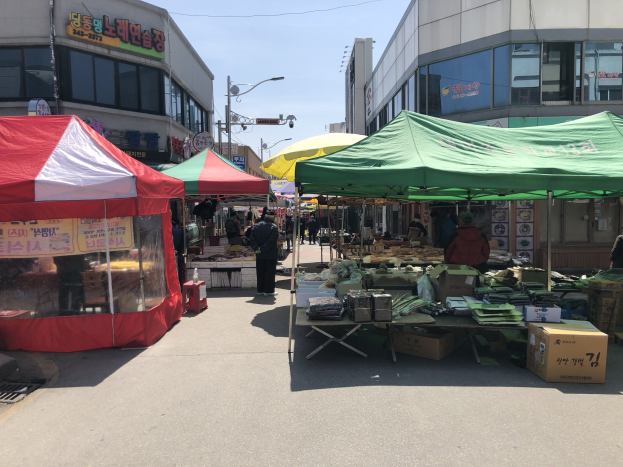 Ein belebter Straßenmarkt in Seoul, Südkorea, mit Zelten, Menschen, Tischen mit Gegenständen, Gebäuden, Laternen und Texttafeln unter einem sichtbaren Himmel.