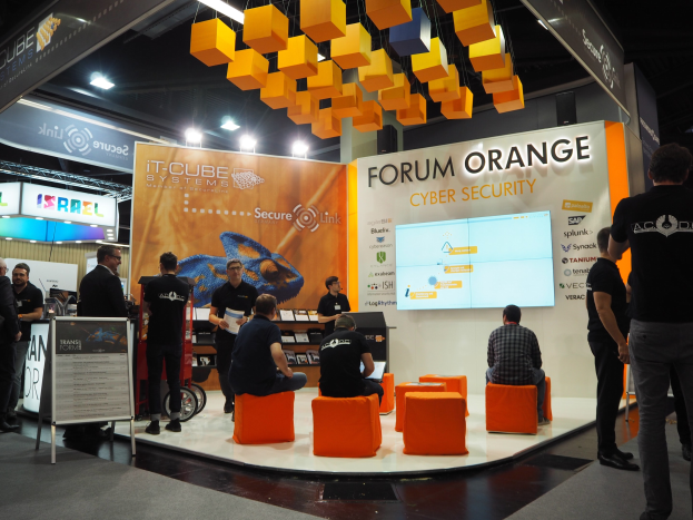 Eine Gruppe von Menschen an einem IT-Cube Systems Forum Orange Cyber Security Stand, mit sitzenden und stehenden Teilnehmern, einer Displaytafel, einem Bildschirm, Regalen, Schildern und Deckenleuchten.