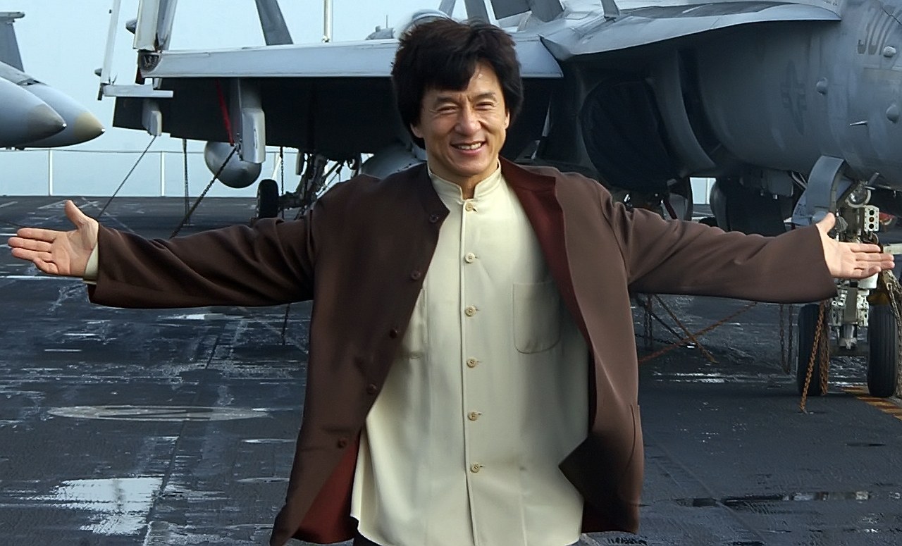 Jackie Chan steht auf dem Deck eines Flugzeugträgers mit ausgestreckten Armen in einem weißen und braunen Kleid, während Kampfflugzeuge und ein klarer blauer Himmel im Hintergrund zu sehen sind.