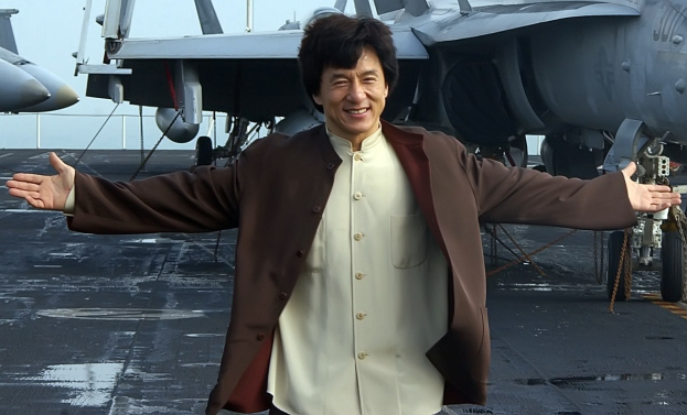 Jackie Chan steht auf dem Deck eines Flugzeugträgers mit ausgestreckten Armen in einem weißen und braunen Kleid, während Kampfflugzeuge und ein klarer blauer Himmel im Hintergrund zu sehen sind.