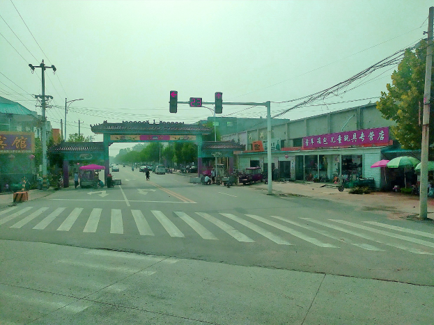 Stadtstraßenkreuzung mit Zebrastreifen, Ampel, Fahrzeugen, Strommasten, Gebäuden mit Beschilderung, Bäumen, Schirmen und einem Bogen im Hintergrund unter einem sichtbaren Himmel.