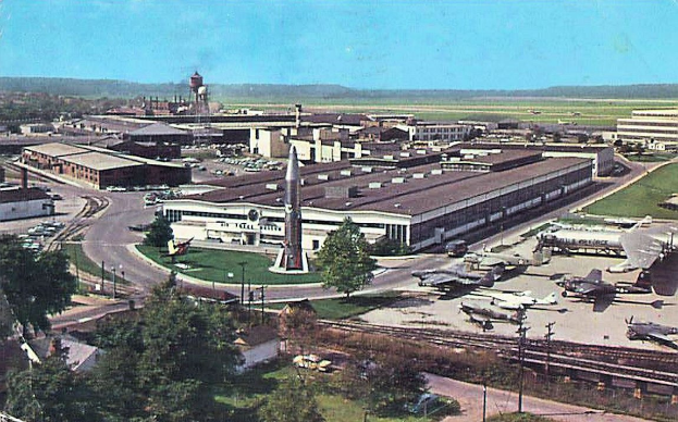 Altes Schwarz-Weiß-Foto eines Flughafens mit parkenden Flugzeugen, Gebäuden, Bäumen, Straßen, Fahrzeugen, Strommasten, Gras, Bahnschienen, Türmen, fernen Hügeln und einem klaren blauen Himmel; ein Text erscheint unten.