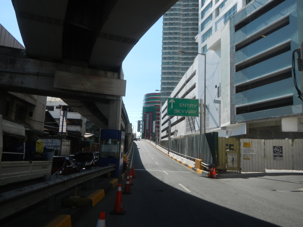 Stadtstraße mit Verkehrskegeln, Fahrzeugen, einer Brücke auf der linken Seite, Gebäuden mit Schildern, Laternenpfählen und einem Himmel als Hintergrund.