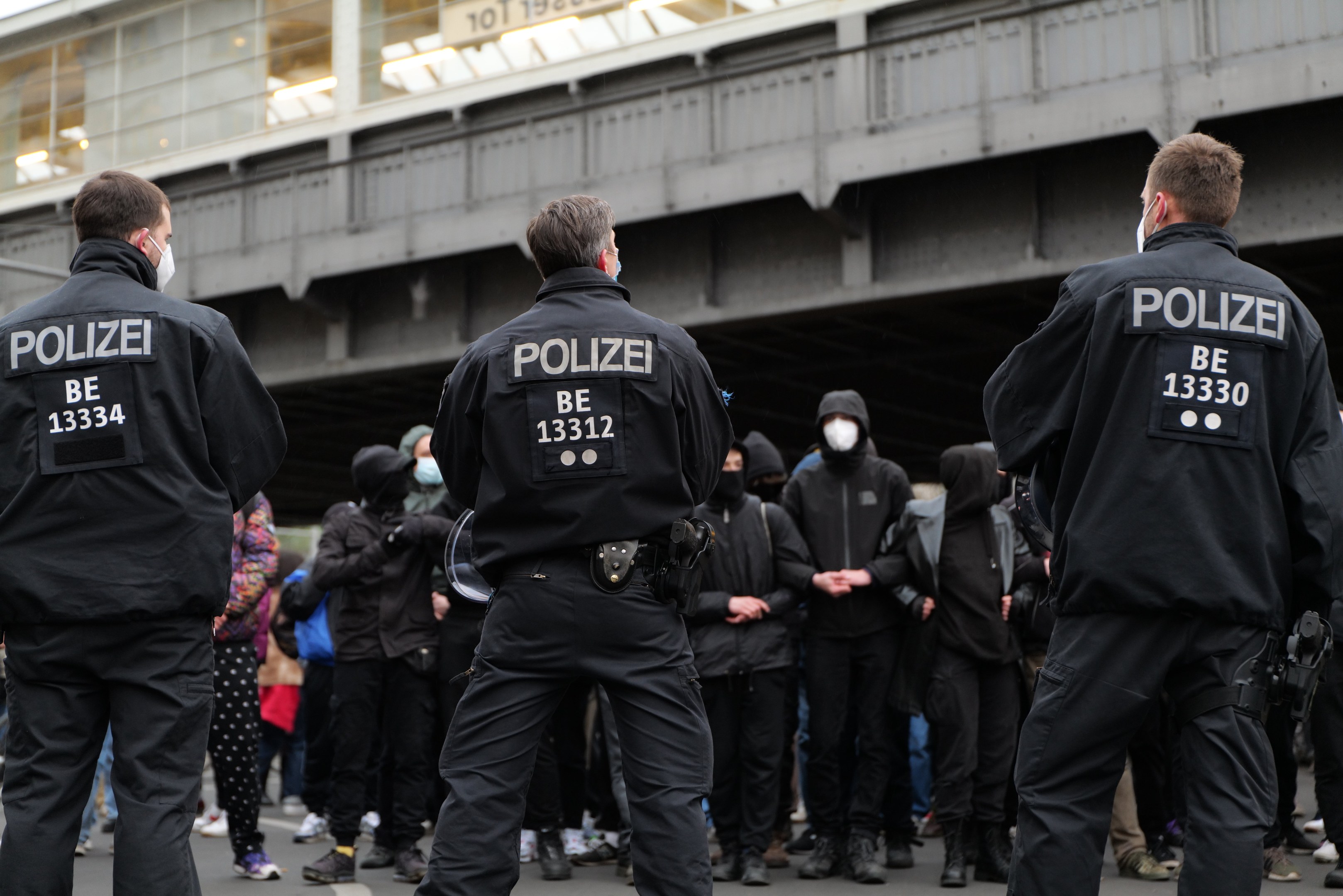Polizeibeamte in schwarzer Uniform und Masken vor einer Menge während einer Demonstration, mit einer Brücke und einem Gebäude im Hintergrund.