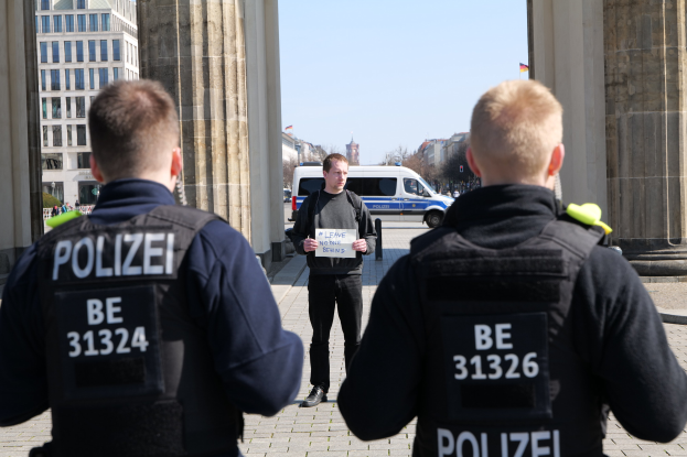 Drei Polizeibeamte stehen vor einem Gebäude, ein Mann in der Mitte hält ein Papier in der Hand, flankiert von Säulen und umgeben von Fahrzeugen, Menschen, Bäumen, Gebäuden und dem Himmel.