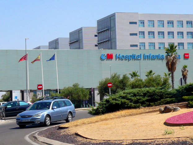 Ein Krankenhausgebäude mit parkenden Autos, umgeben von städtischen Elementen wie Gebäuden, Straßenmasten und Fahrzeugen, unter einem klaren blauen Himmel.