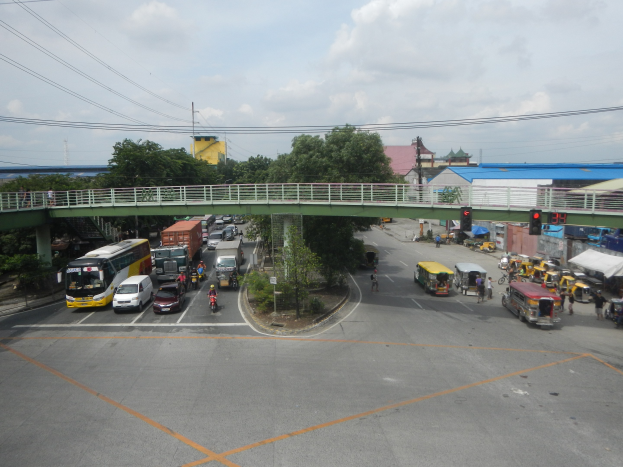 Eine belebte Stadtstraße mit Autos, Bussen, Fußgängern auf einem Gehweg, einer Brücke mit Geländer, Verkehrsampeln, Bäumen, Gebäuden, Strommasten mit Kabeln und einem bewölkten Himmel.