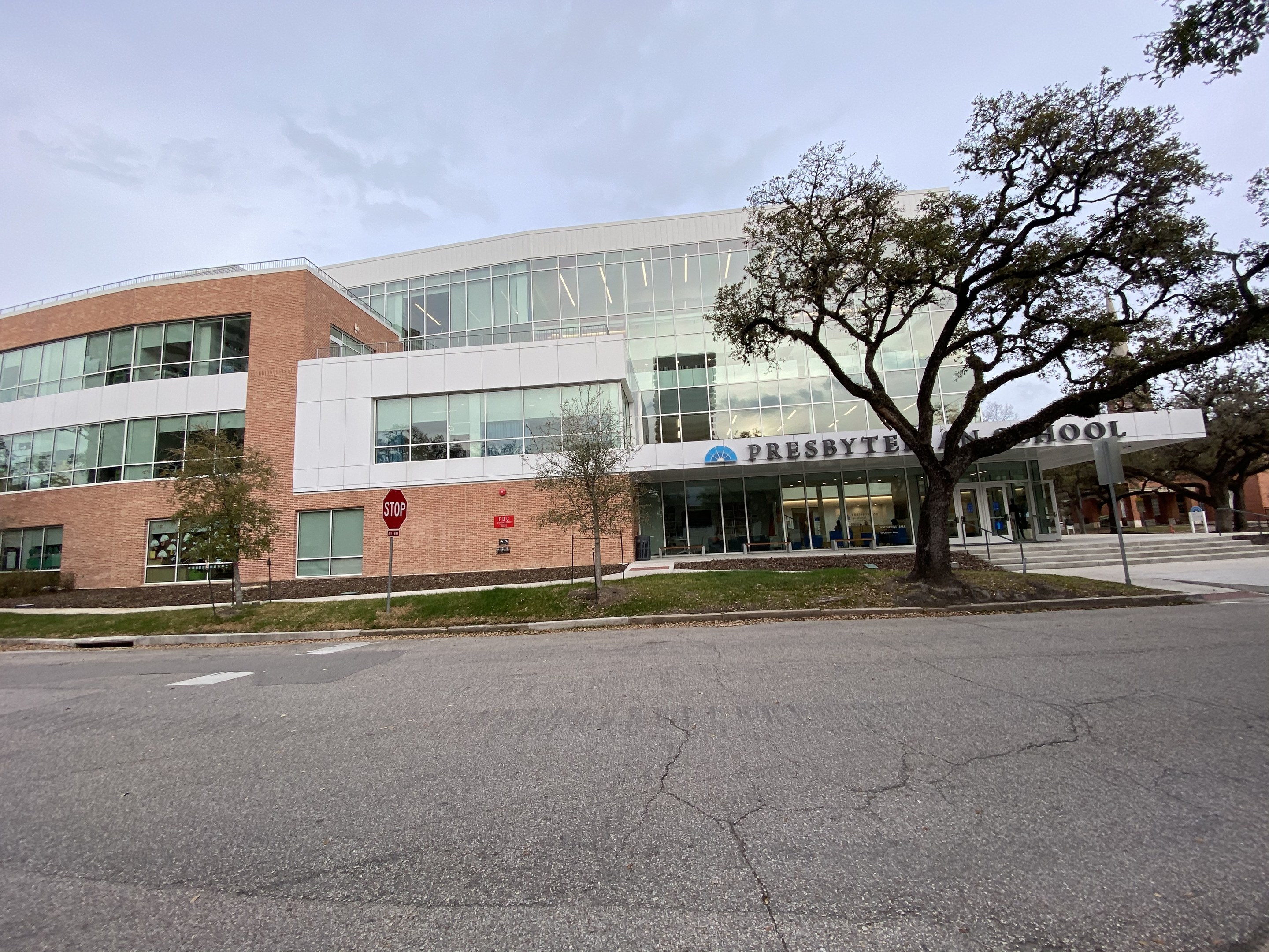 Großes zweistöckiges Backsteingebäude mit der Aufschrift "Presbyte Mittelschule" und vielen Fenstern, umgeben von Bäumen, Gras, Schildern, Strommasten und einer Straße unter einem bewölkten Himmel.