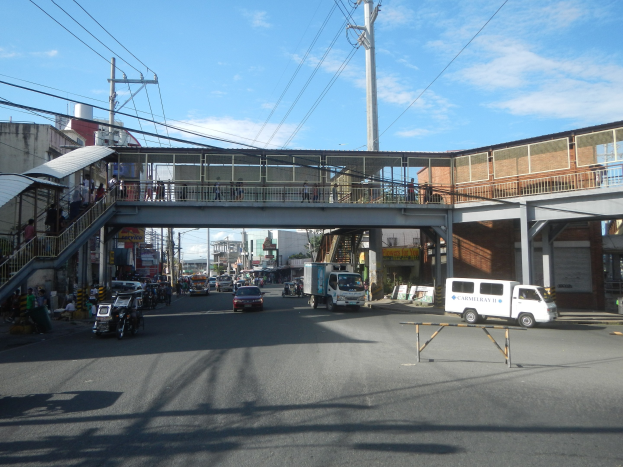 Eine Stadtstraße mit fahrenden Autos, eine Fußgängerbrücke mit Menschen, Strommasten mit Drähten, Gebäude mit Fenstern und ein bewölkter Himmel.