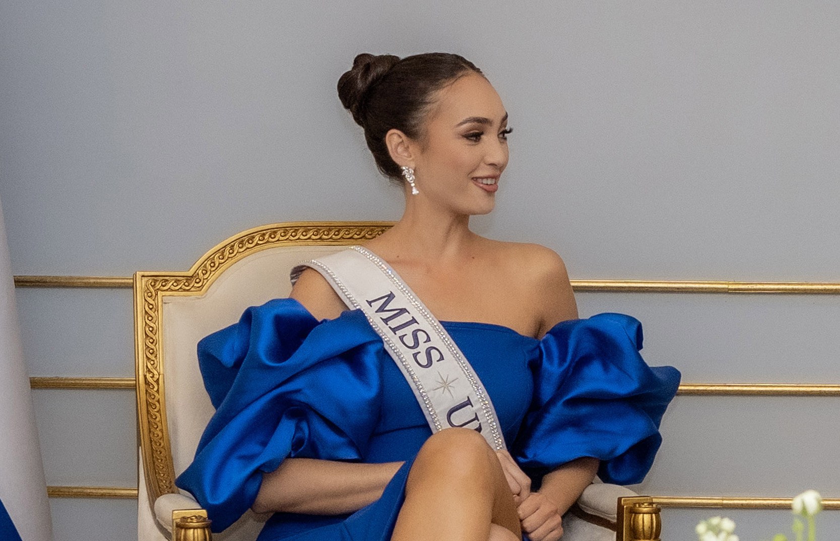 Eine Frau in einem blauen Kleid trägt einen "Miss Universe 2019"-Schal und sitzt lächelnd auf einem Stuhl umgeben von Blumen mit einer Wand im Hintergrund.