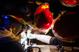 Feuerwehrleute in Helmen und Handschuhen arbeiten an einem Feuerwehrauto in einer dunklen Umgebung, mit einem Tisch voller Gegenstände rechts daneben.