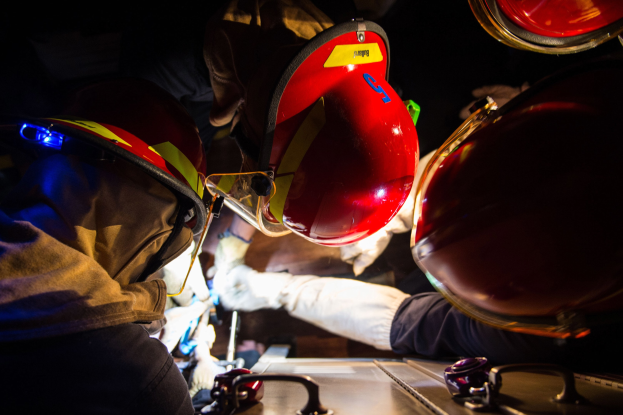 Feuerwehrleute in Helmen und Handschuhen arbeiten an einem Feuerwehrauto in einer dunklen Umgebung, mit einem Tisch voller Gegenstände rechts daneben.