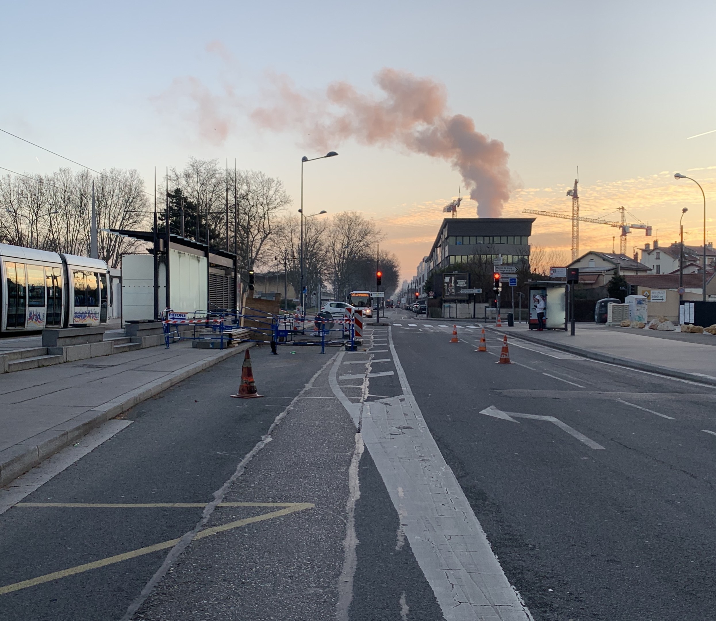 Eine Stadtstraße mit Verkehrskegeln, einem Bus, Laternenmasten, Bäumen, Gebäuden, Kränen und Rauch, der aus einem fernen Gebäude aufsteigt.