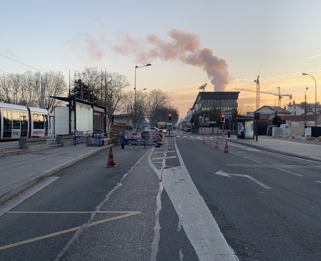 Eine Stadtstraße mit Verkehrskegeln, einem Bus, Laternenmasten, Bäumen, Gebäuden, Kränen und Rauch, der aus einem fernen Gebäude aufsteigt.