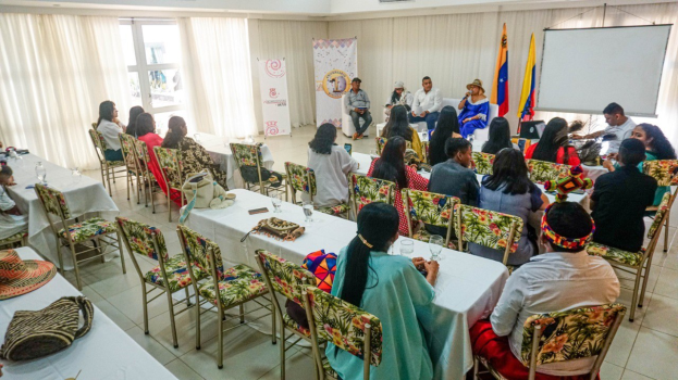 Gruppe von Menschen an Tischen in einem Raum mit Gegenständen auf den Tischen, Flaggen und Bannern im Hintergrund und Deckenleuchten, was auf eine indigene Kulturwerkstatt hinweist.
