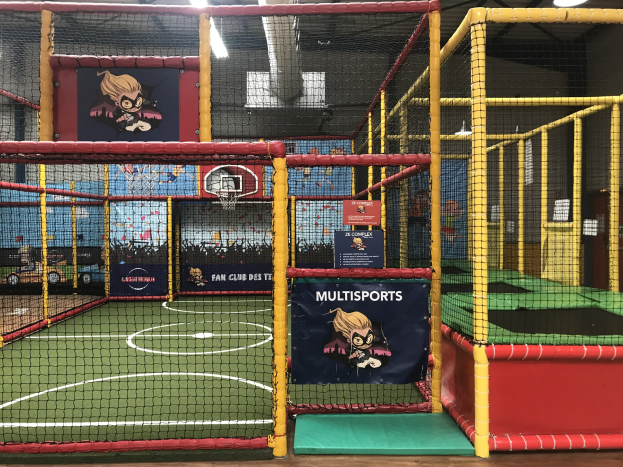 Großes Indoor-Trampolinpark mit einem Basketballfeld in der Mitte, umgeben von einem Netz, mit einem Basketballkorb, Deckenleuchten und Wandbannern.
