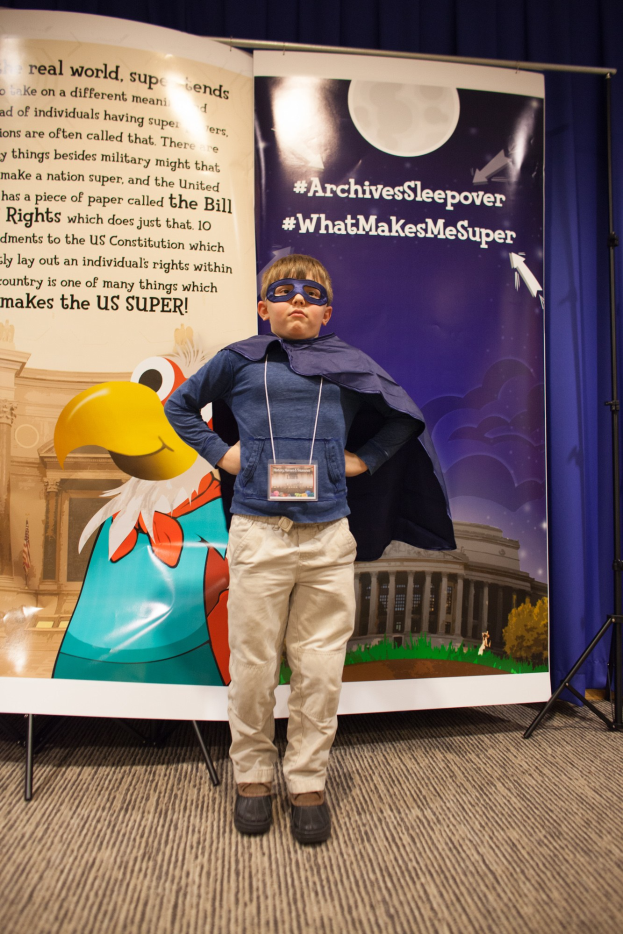 Ein junger Junge in einem Superheldenkostüm steht vor einem Banner mit Text und einem Vogelbild, mit einem Vorhang im Hintergrund und Schuhen.