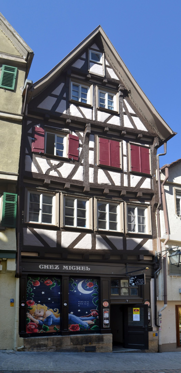 Altes Gebäude mit roten Fensterläden, mehreren Fenstern und klassischem Design, das sich auf einer Straße unter einem klaren blauen Himmel befindet.