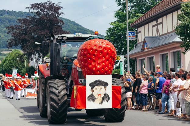 Ein Traktor mit einer großen Erdbeere auf dem Rücken fährt eine Straße entlang, während eine Gruppe von Menschen mit Fahrrädern auf dem Bürgersteig steht, mit Gebäuden, Bäumen und Hügeln im Hintergrund unter einem klaren blauen Himmel.