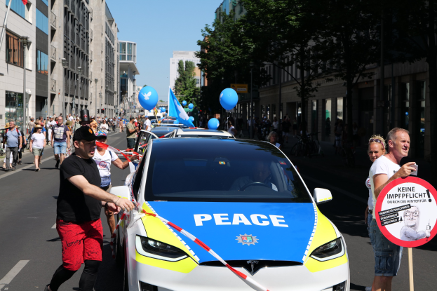 Eine Gruppe von Menschen geht neben einem Polizeiwagen her, der mit blauen, gelben und roten Schleifen geschmückt ist und auf dem das Wort "Frieden" zu sehen ist, vor einer Kulisse aus Gebäuden, Bäumen und einem klaren blauen Himmel.