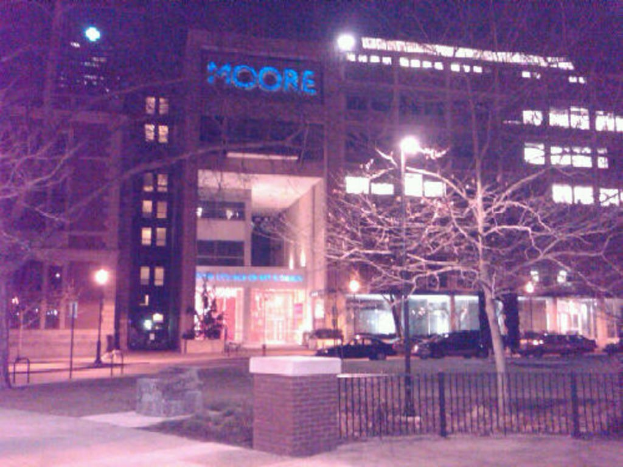 Ein Krankenhausgebäude mit parkenden Fahrzeugen, Bäumen und einem Zaun davor.