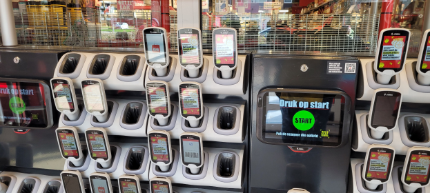 Ein Verkaufsautomat mit mehreren Handys auf der Oberseite, vor einem Hintergrund aus Menschen, Geschäften, Schildern und anderen urbanen Elementen.