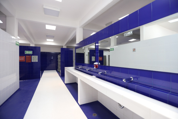 Öffentliche Toilette mit blauen Kachelwänden, weißen Countertops, Waschbecken mit Hähnen, einer Plattform mit einer Flasche und anderen Gegenständen, Texttafeln an den Wänden, Deckenleuchten und Pfeilern.