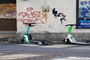Zwei Elektro-Scooter nebeneinander auf einer Stra√e geparkt, mit einer Graffiti-bedeckten Wand und einem Plakat an einem Fenster im Hintergrund.