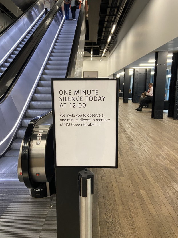 Eine Rolltreppe in einem Flughafen mit einem Schild, auf dem "Eine Minute Stille heute" steht, einige Menschen darauf und an der Decke befestigte Lampen im Hintergrund.