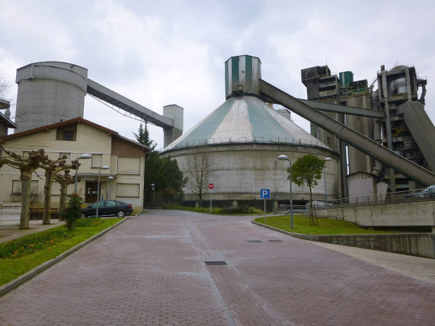 Große Zementfabrik mit Gebäuden, Straßeninfrastruktur, Fahrzeugen und Grünflächen gegen einen bewölkten Himmel.